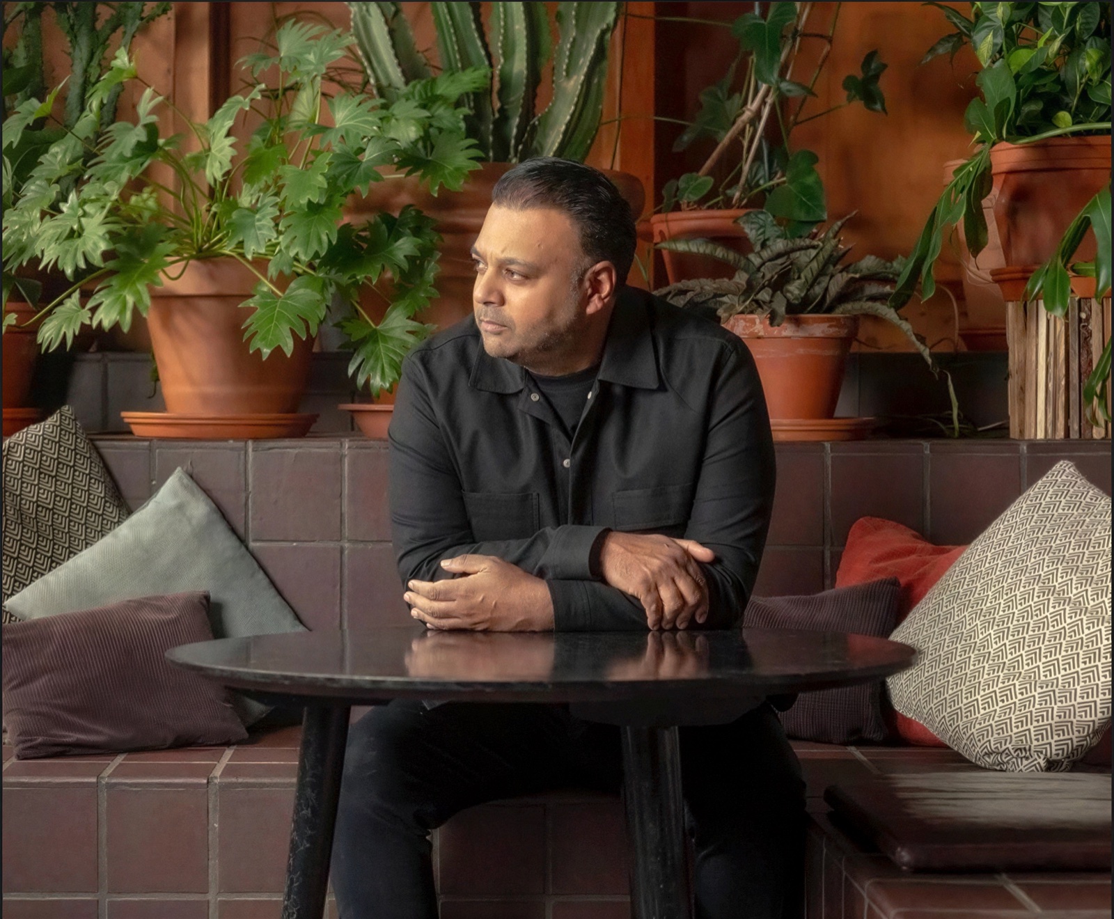 Taz Khan MBE, founder of London's Community Kitchen, seated at a table surrounded by potted plants.